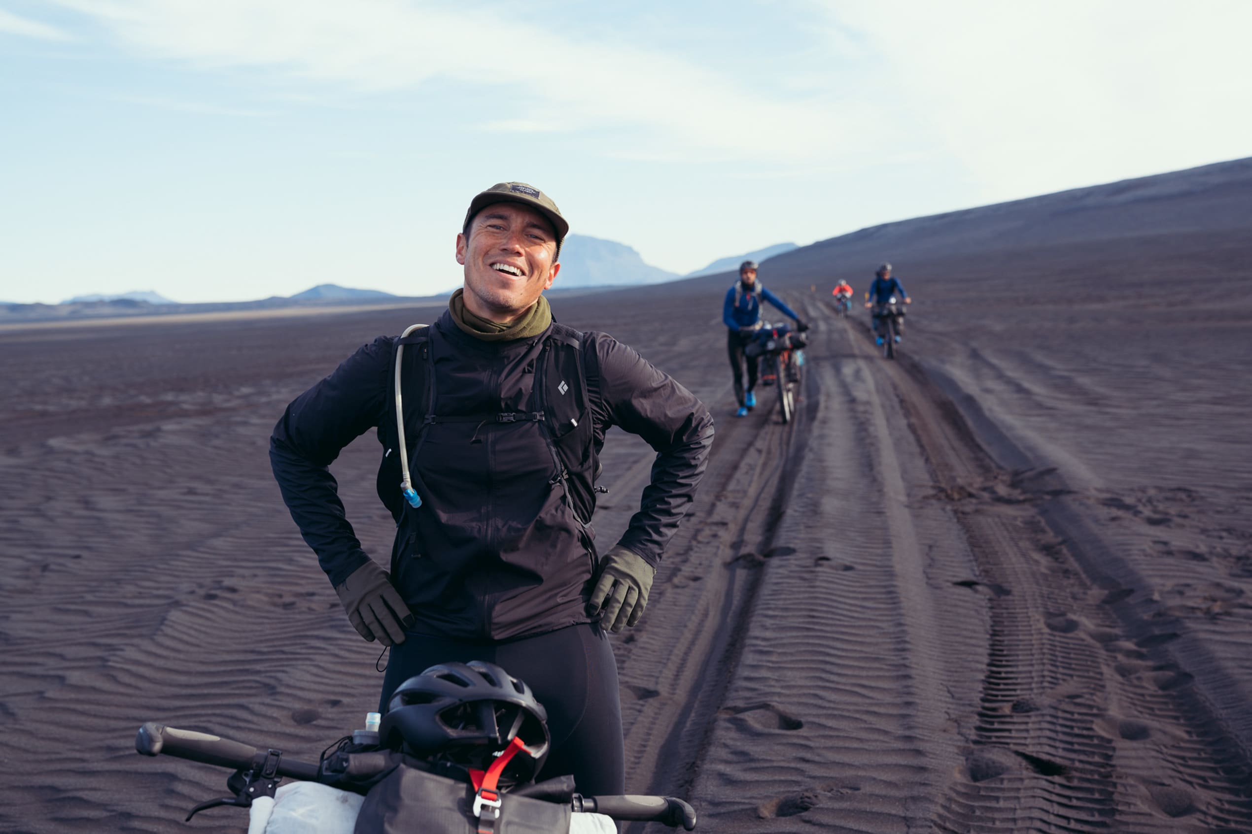  Fjórir hjólreiðakappar á svörtum sandi, með fjöll í bakgrunni. Einn er í forgrunni og hinir þrír í bakgrunni.