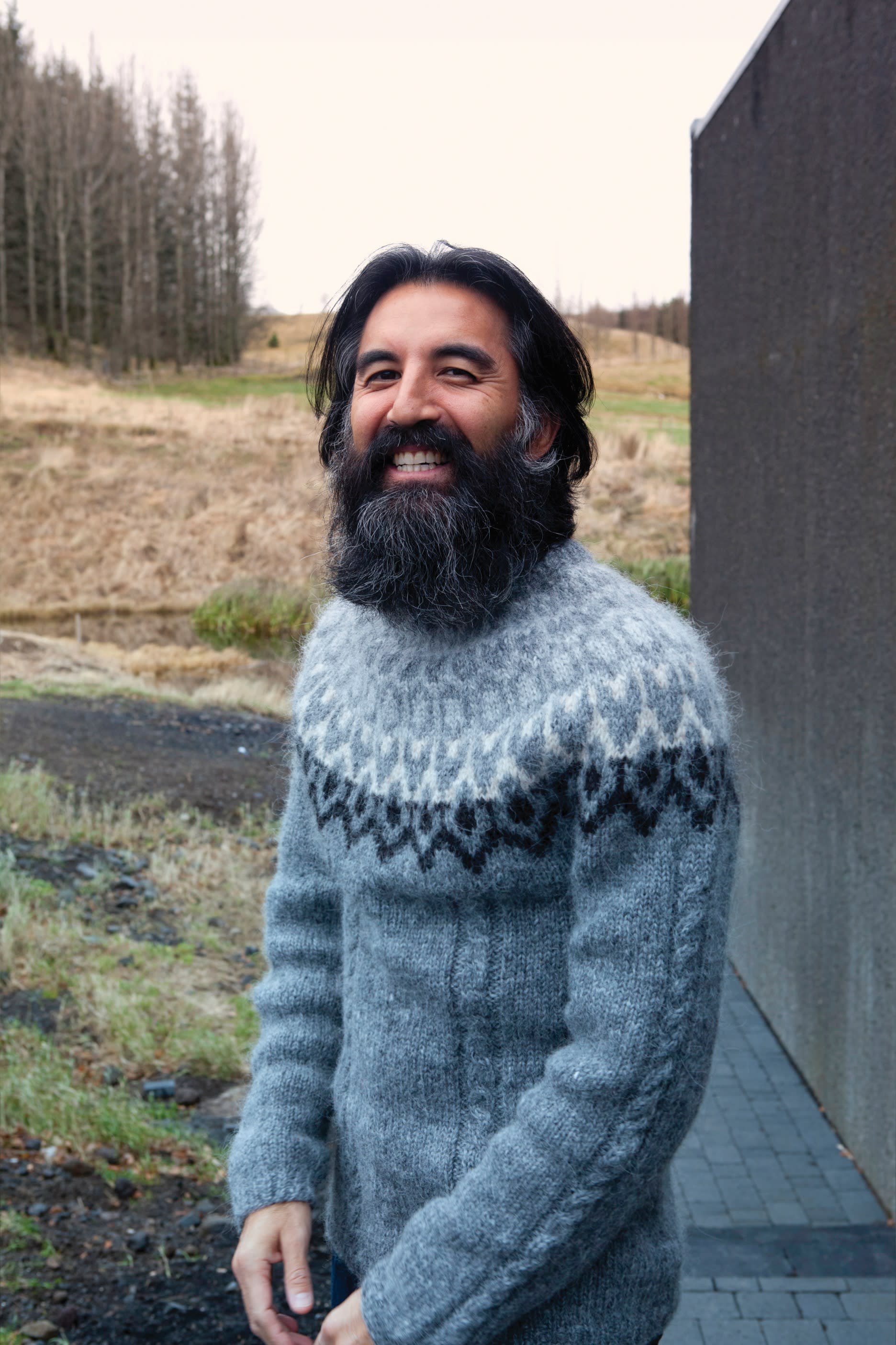 a man with a large beard, standing in the Icelandic nature, smiling directly into the camera while wearing a grey knitted Icelandic sweater