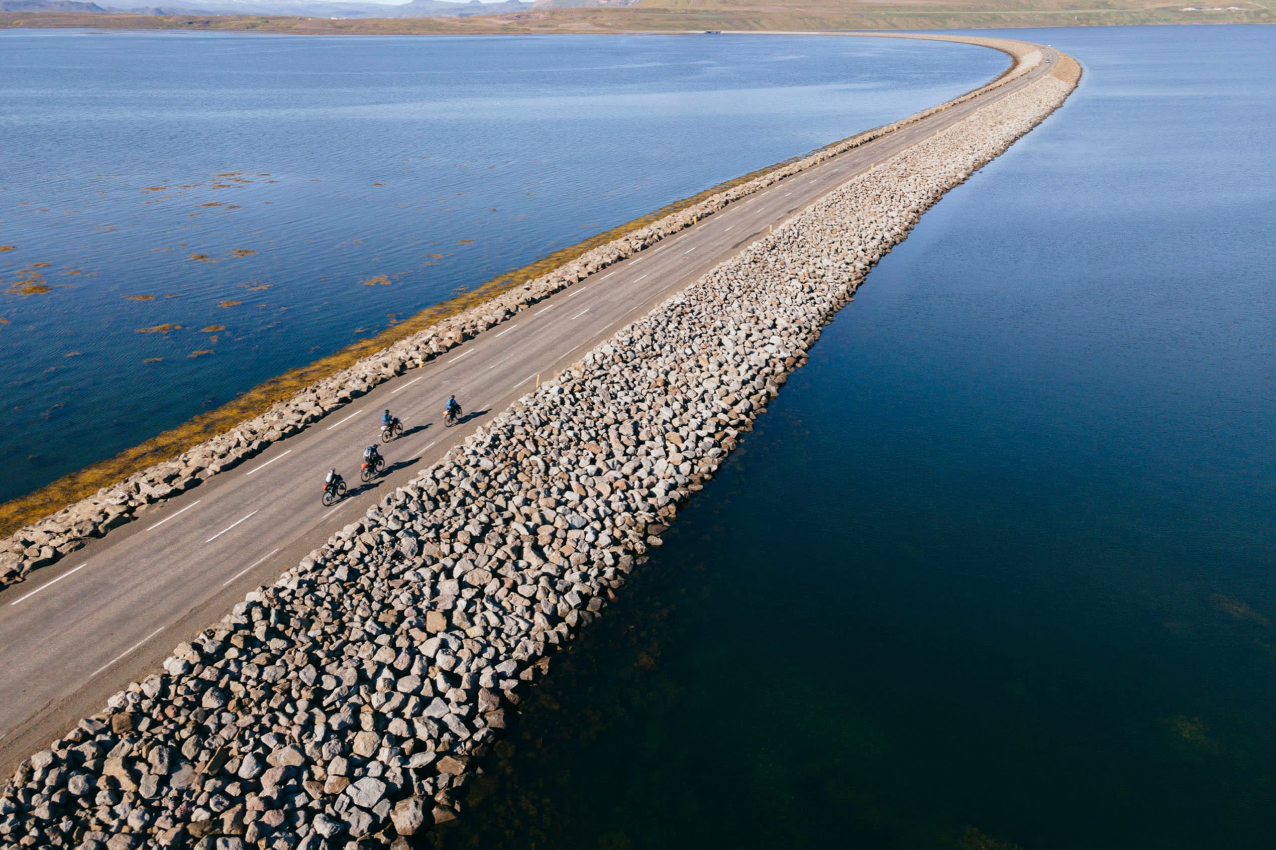 Four cyclists riding through a road which leads through a body of relatively still water