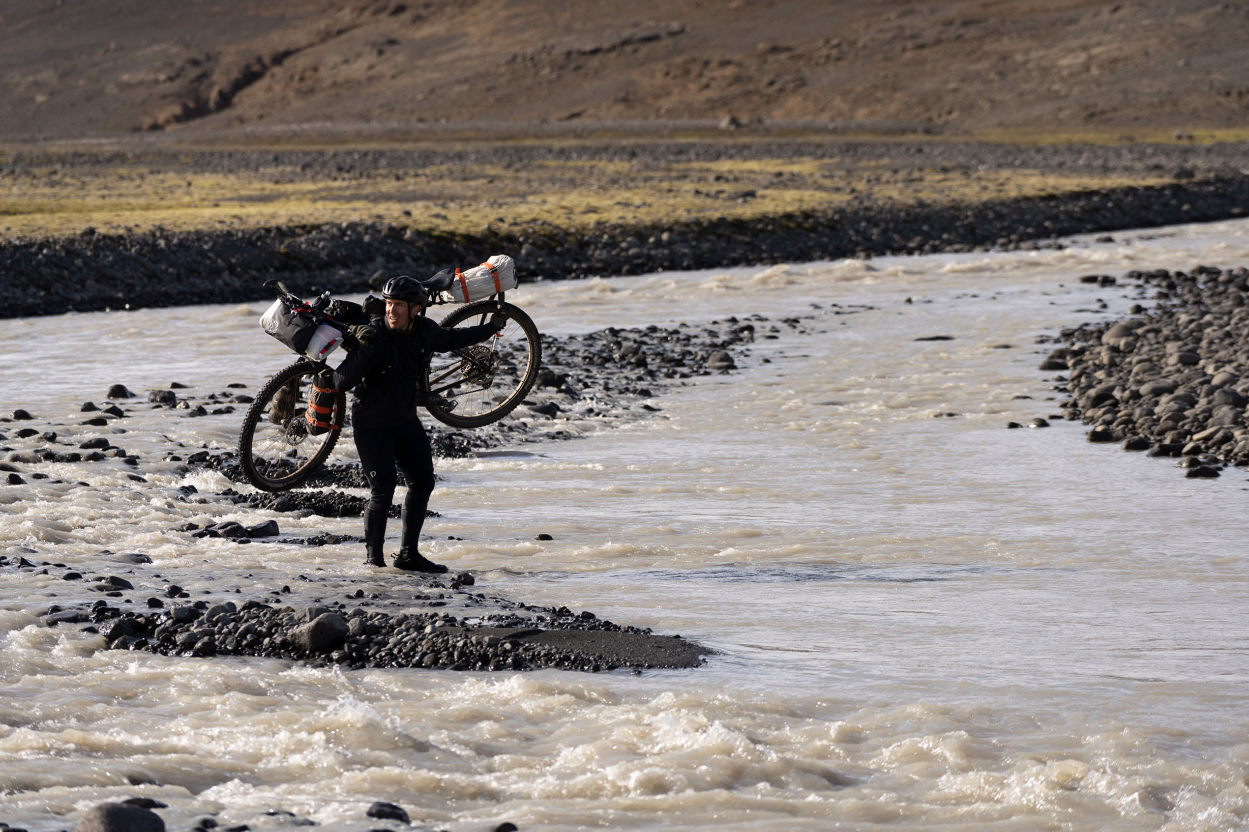 an image of a man carrying a bike over his shoulders and walking through a river in Iceland