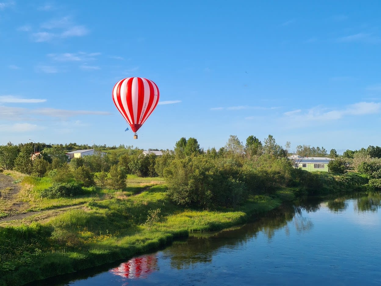 a red and white striped hot air balloon flying over the leafy green Icelandic landscape next to a body of water