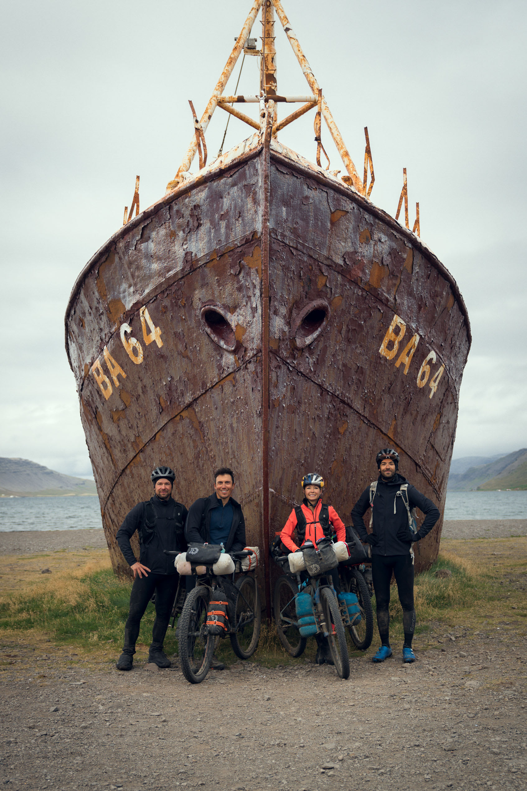 Fjórir hjólreiðakappar stilltir upp við strandað og ryðgað skip, BA 64. Í bakgrunni sjór, himinn og fjall.