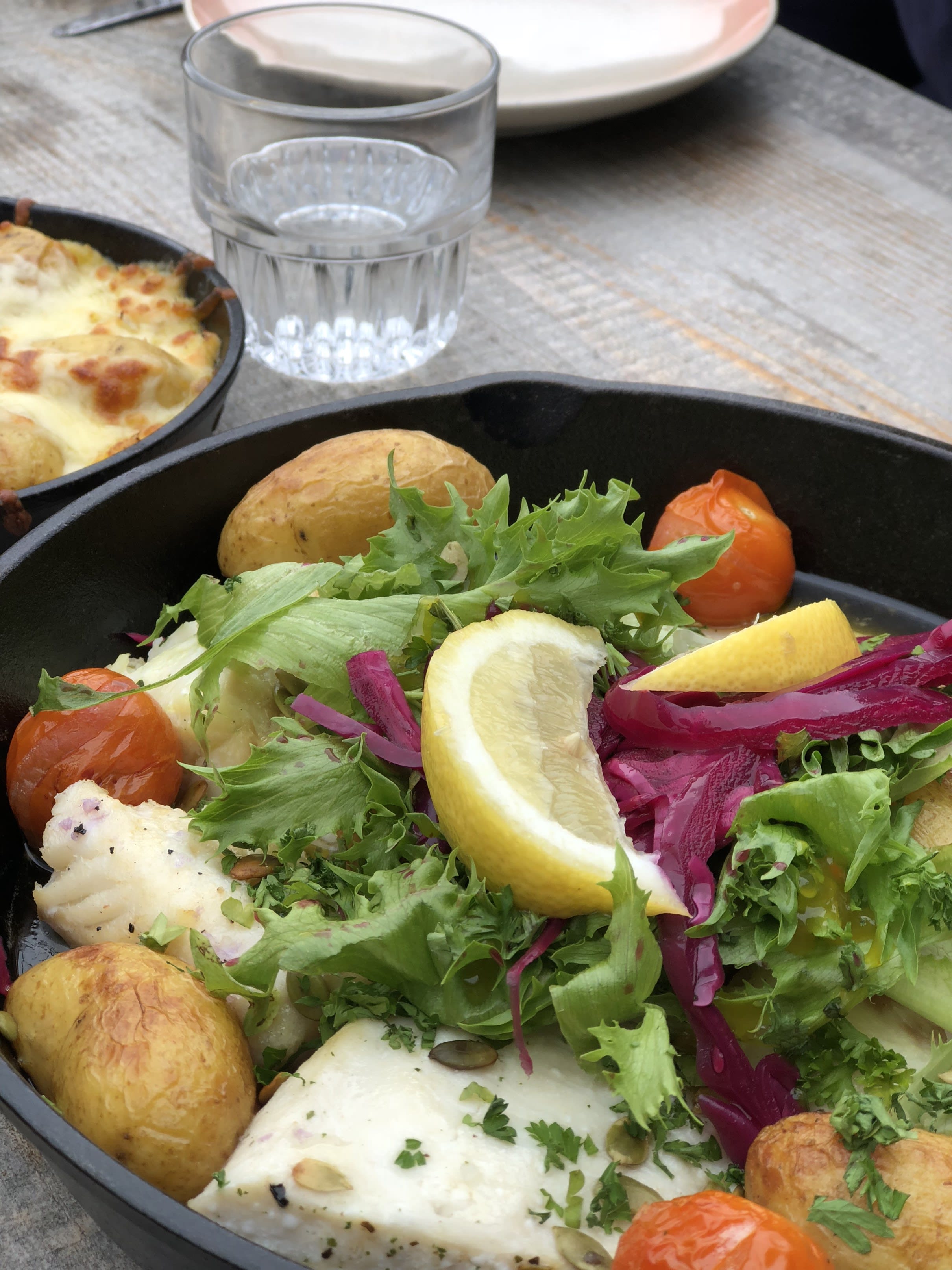 a close up of a dish with fish, salad, potatoes, cabbage and lemon in a black oven dish