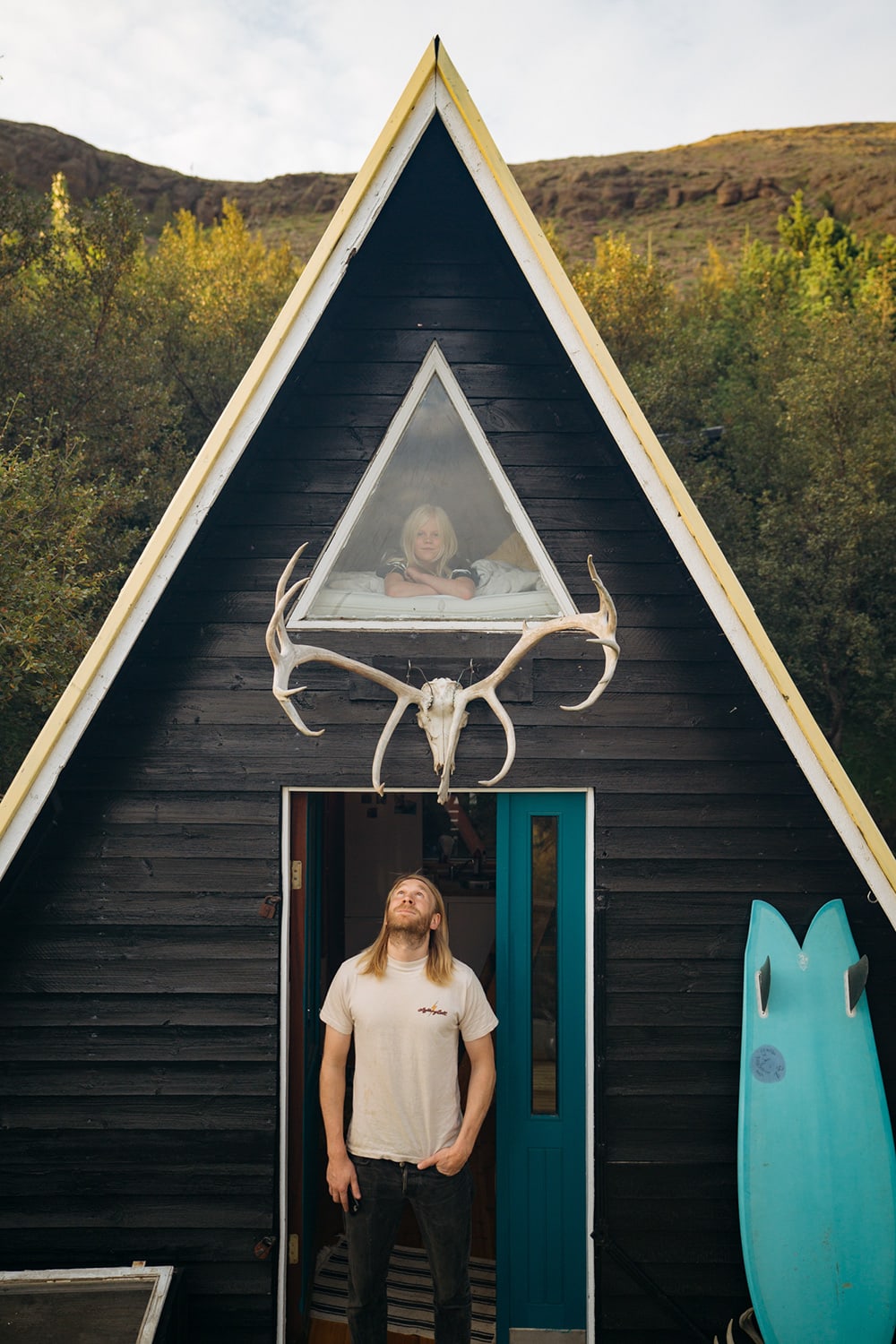 an adult standing in the doorway to a small cabin looking upwards at a child in the window above