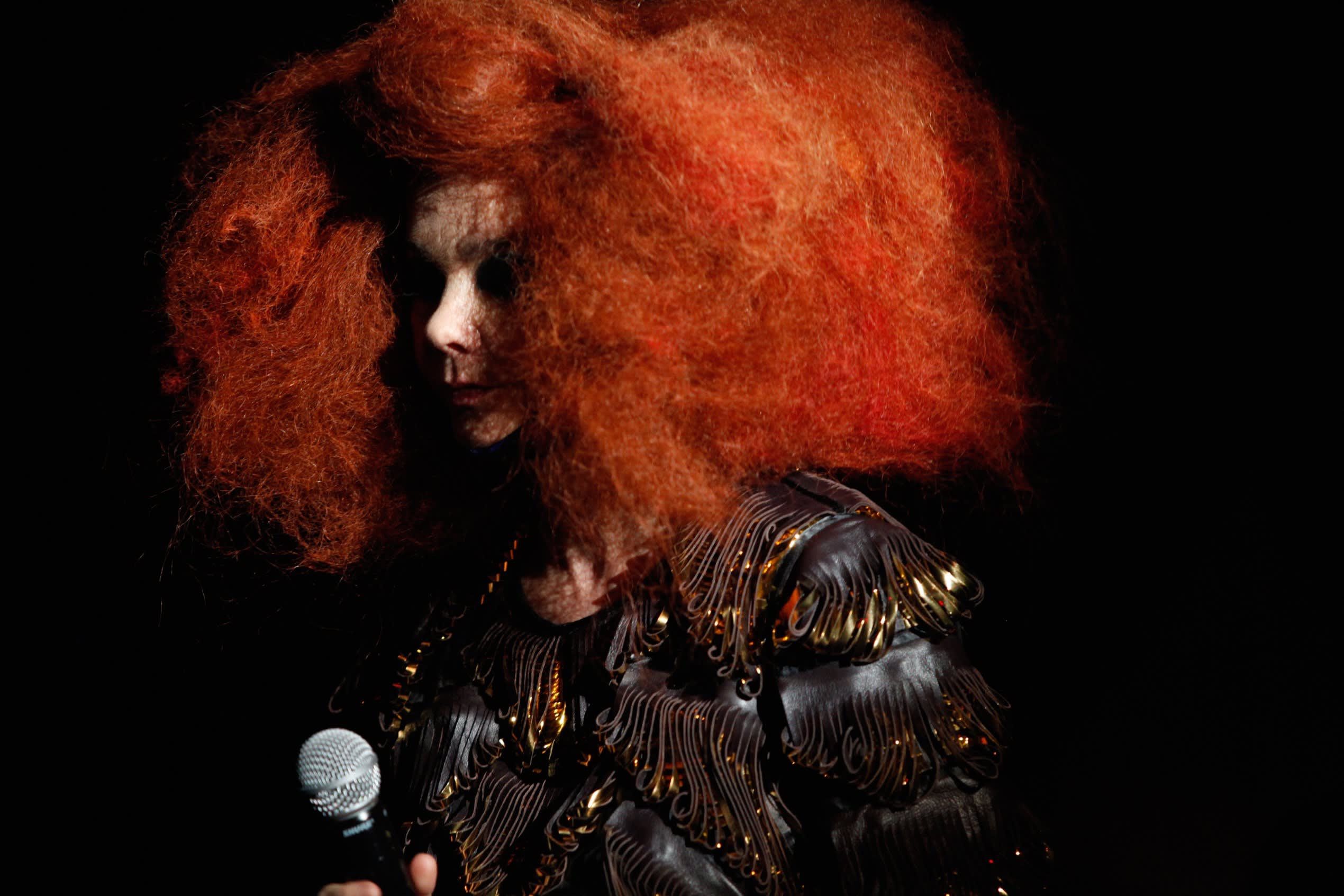 bjork pictured with a bright red hair style, wearing a black and gold jacket and facing side-on to the camera in a dark room