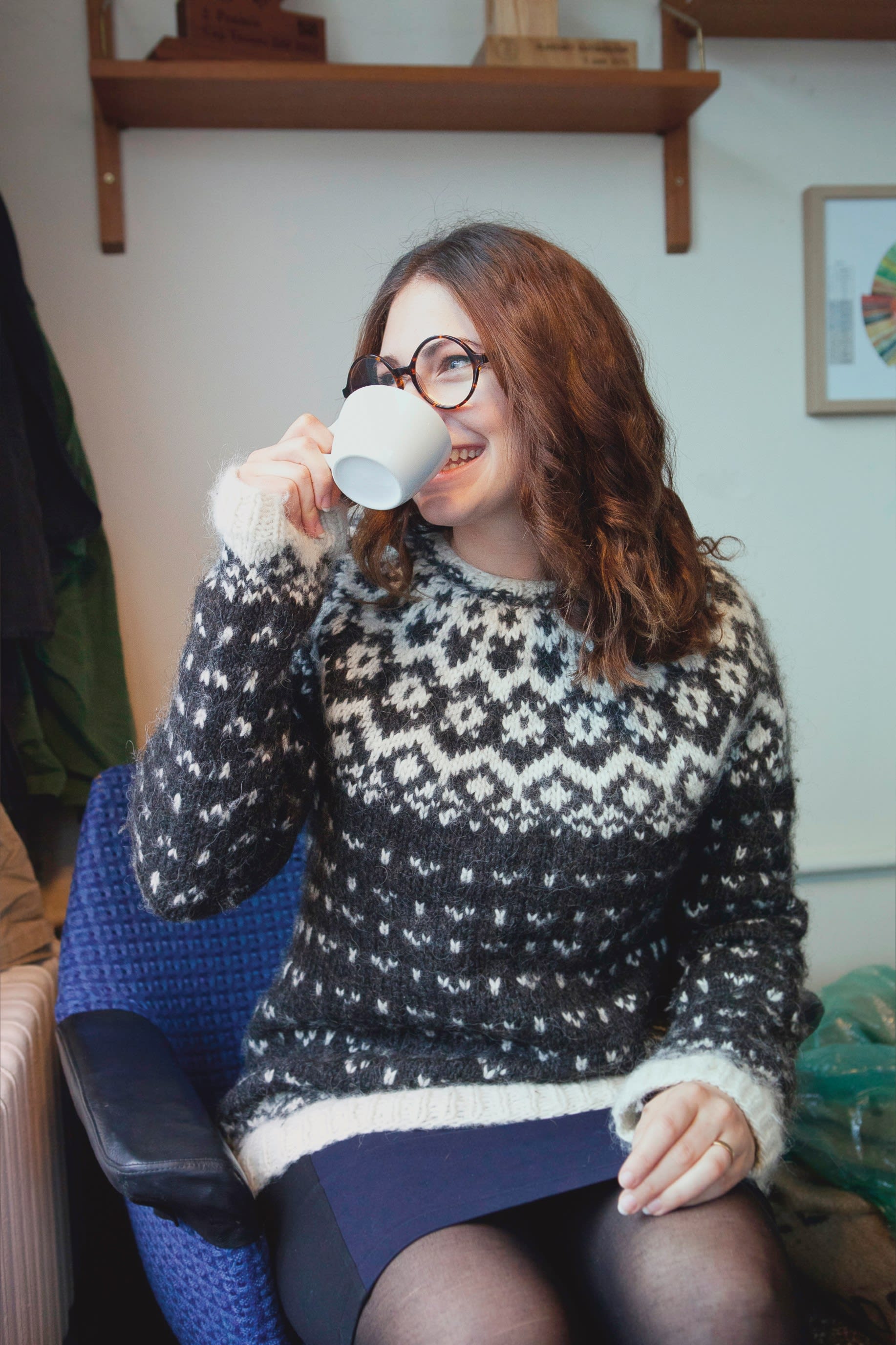 a woman with brown curly hair drinking from a white cup in a cafe and wearing a black and white knitted Icelandic sweater