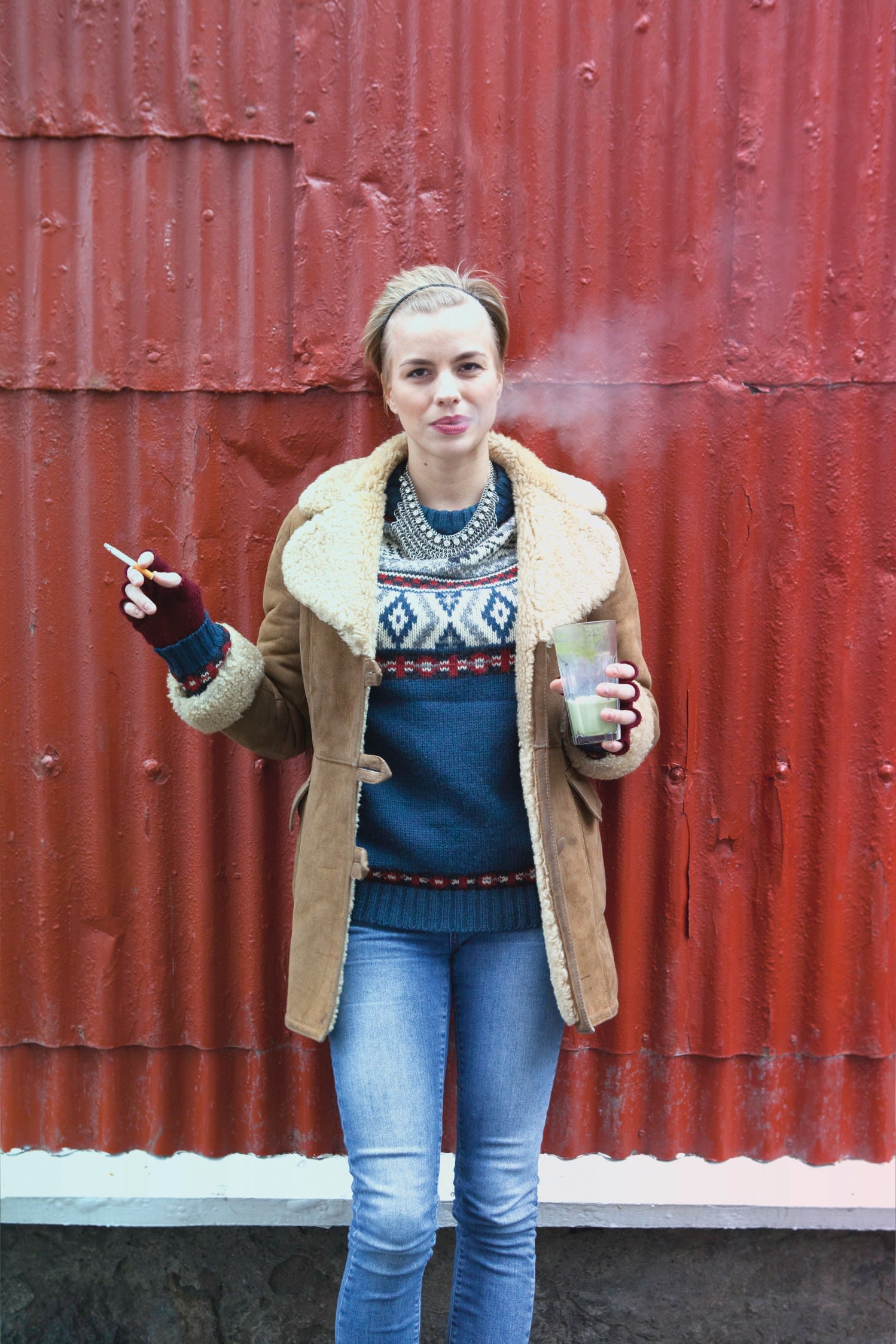 a woman standing against a red corrugated metal building wearing a blue Icelandic sweater with a green drink in one hand and a cigarette in the other