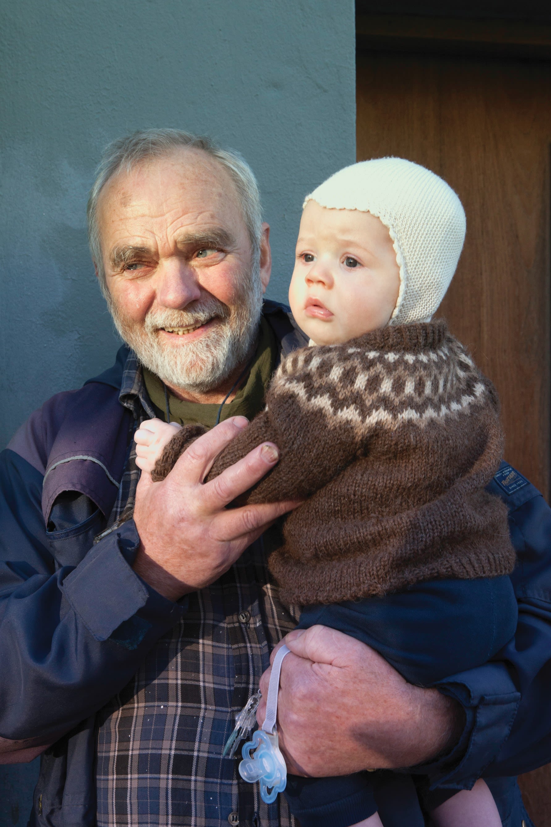 an older man holding a small baby who is wearing a brown knitted Icelandic wool sweater