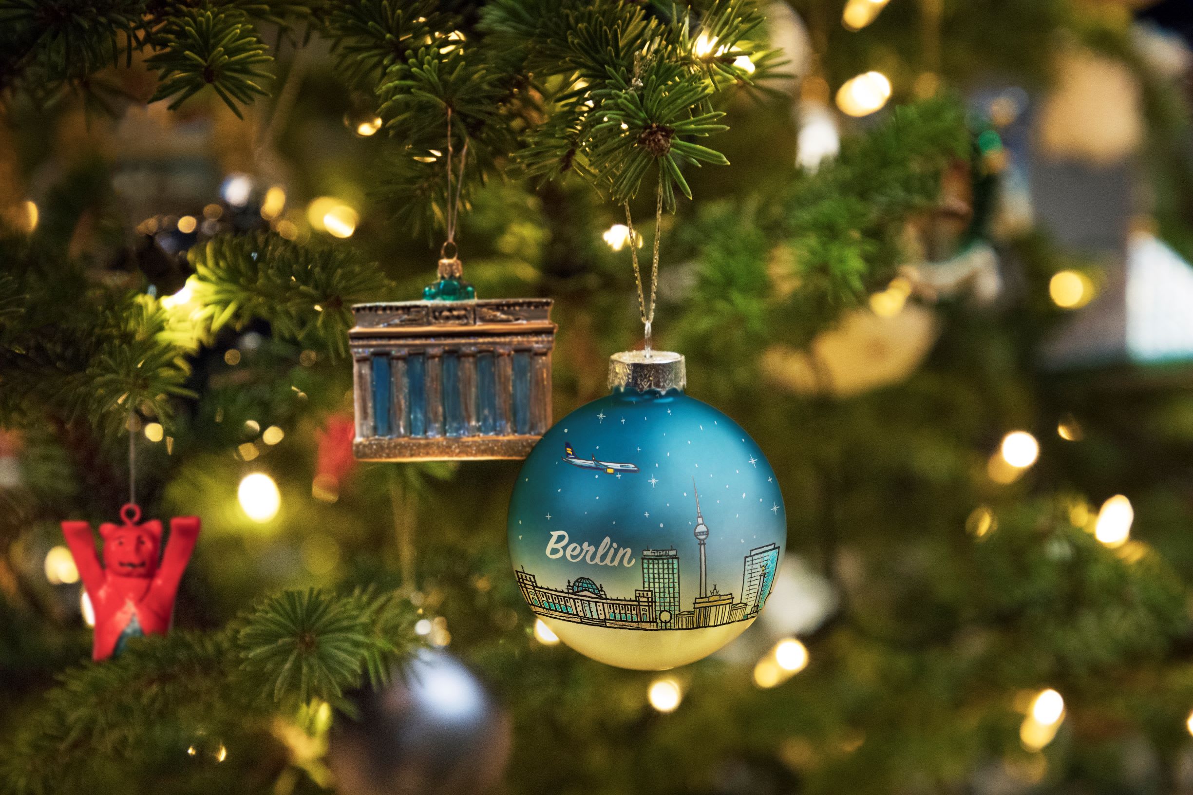 A blue bauble which reads 'Berlin' and has a city scape scene, positioned next to a decoration of the Brandenberg gate, both hanging from a green Christmas tree