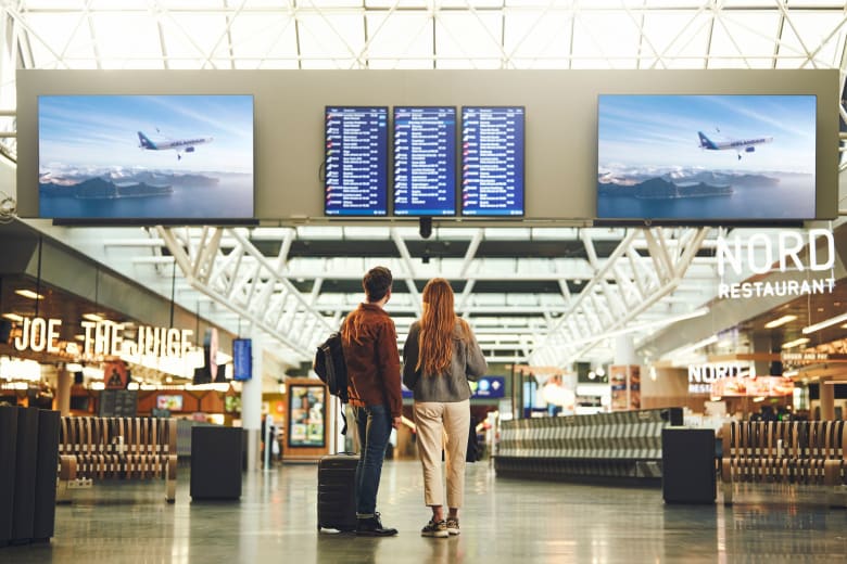 A couple with carry-on baggage looking at a flight schedule screen at Keflavík International Airport