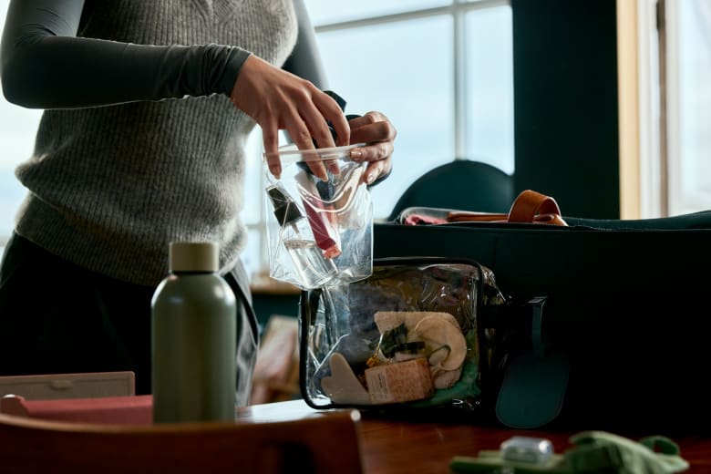 A person is packing a transparent toiletry bag with various items, including a toothbrush and other personal care products, on a wooden table. A water bottle and an open travel bag are also visible.